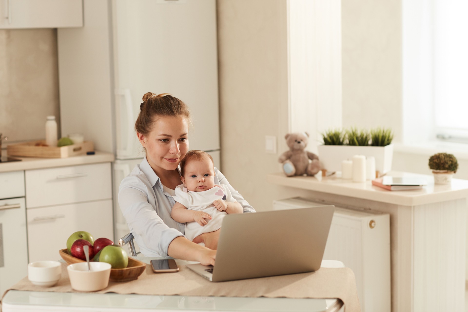 Young smiling mother in blue shirt sitting in kitchen and trying to entertain babt by showing cartoons on laptop
